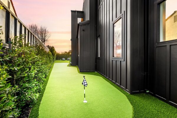 A modern backyard with a mini-golf putting green next to a black contemporary house at sunset.